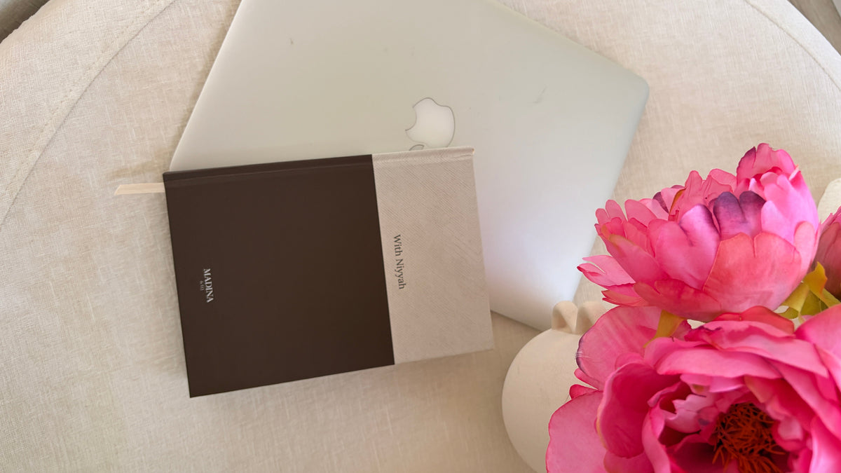 A minimalist hardcover notebook with a beige top half and dark brown bottom half, placed on a light beige fabric surface next to a closed silver laptop and pink flowers in a white vase.